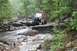 1984 Ford Bronco II - A person stands next to a modified off-road vehicle as it navigates a challenging rocky terrain with a stream running through it.