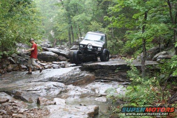 1984 Ford Bronco II off-roading over rocks with a person walking nearby