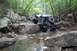 1984 Ford Bronco II - A heavily modified off-road vehicle carefully maneuvers through a shallow, rocky stream bed surrounded by dense woods.