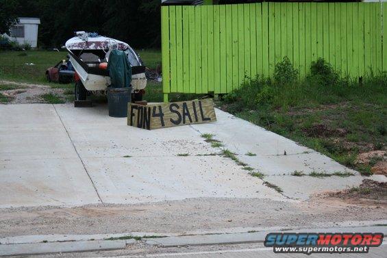 Boat on trailer with 'FUN 4 SAIL' sign in front, next to a green fence