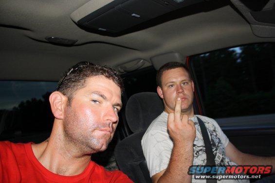 Guy in red shirt and another guy giving the finger while riding in a car