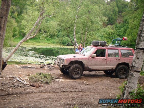 Red 1998 Jeep Cherokee parked off-road near a lake, with a spare tire on the roof rack