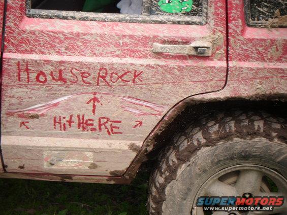 Mud-covered red 1998 Jeep Cherokee side with graffiti reading 'House Rock' and 'Hit HERE'
