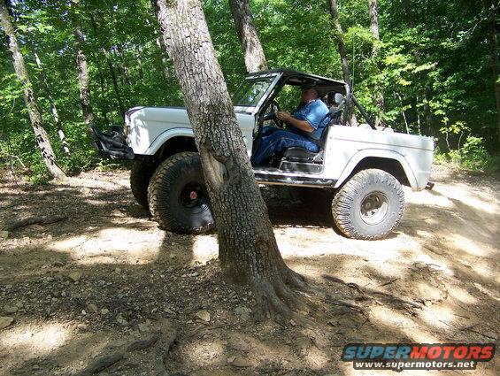 White off-road vehicle with large tires, driven by a man, on a dirt path in a wooded area