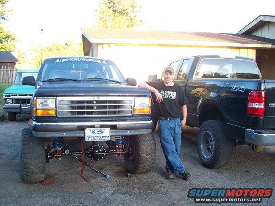 Black Ford Explorer lifted with large tires, man in graphic t-shirt leaning on it. Green Ford truck behind