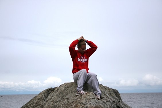 Person in red sweatshirt sitting on a rock by the water, hands in hair