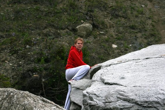 Woman in red hoodie and grey pants climbing large grey rocks with green foliage in background