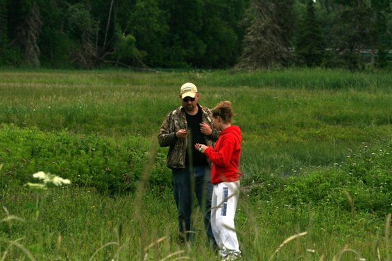 Man in camouflage jacket and woman in red hoodie stand in a field of tall grass