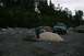 1990 Chevrolet Suburban - A person works near a modified light blue SUV on a rocky shoreline with trees in the background.