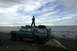 1990 Chevrolet Suburban - A person stands atop a modified SUV parked on a rocky beach with the ocean stretching to the horizon under a cloudy sky.