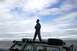 1990 Chevrolet Suburban - A person stands on the roof rack of a gray SUV parked near the ocean under a cloudy sky.
