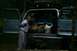 1990 Chevrolet Suburban - A young woman gestures towards the packed cargo area of a light blue SUV, its rear doors open.