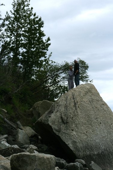 Two people stand on a large rock formation near trees and a cloudy sky