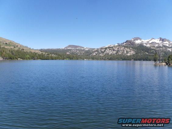 Lake with mountains and trees under a clear blue sky. "SUPERMOTORS www.supermotors.net" logo in lower right