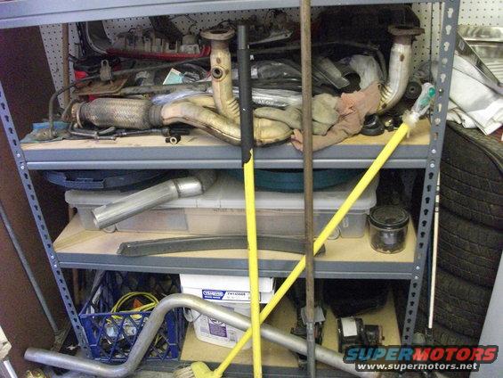 Shelving unit filled with various automotive exhaust pipes and mufflers, some tools leaning against it
