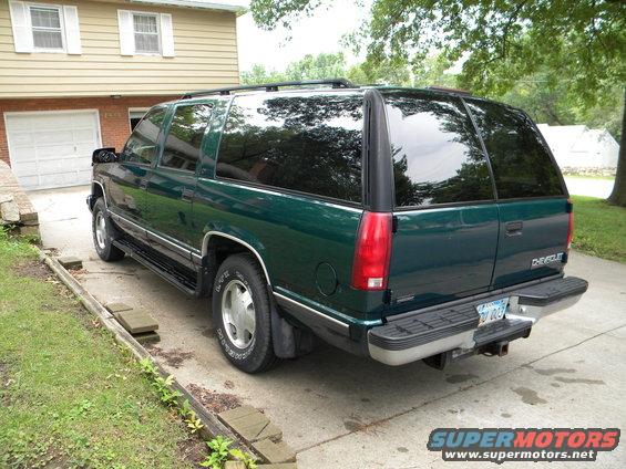 Green 1999 Chevrolet Suburban parked in driveway, rear 3/4 view. Tinted windows, chrome bumper, license plate visible
