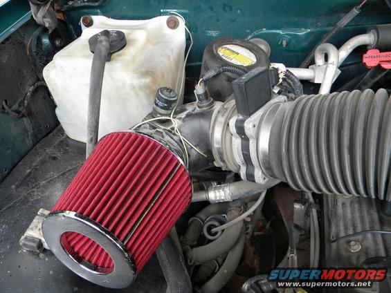 Engine bay with green paint, white coolant reservoir, and red cone air filter. A black sensor is attached to the intake
