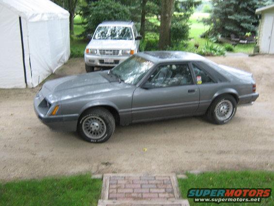 Gray 1985 Ford Mustang parked outside next to a white tent and an SUV