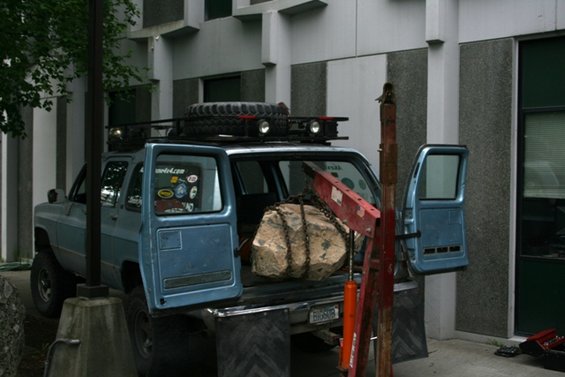 Blue SUV with open doors, roof rack, spare tire, and a large rock chained in the bed
