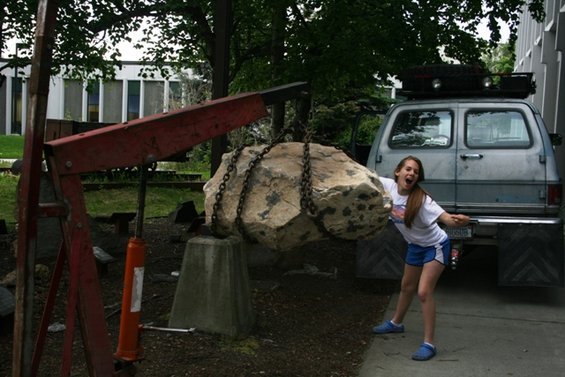 Girl posing with a large rock being lifted by a red crane next to a blue van