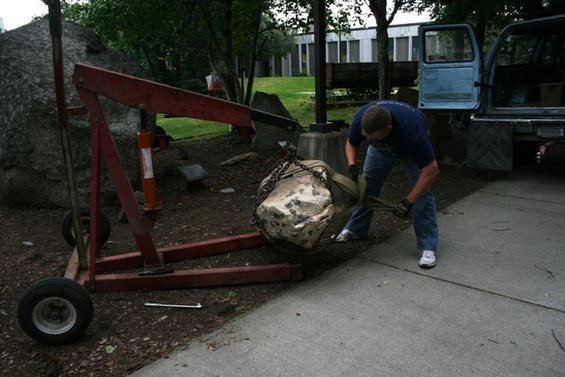 Engine hoist lifting a large rock with a man securing straps. Blue truck in background