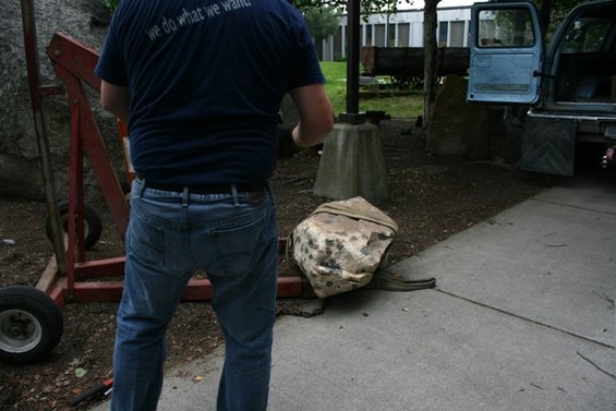 Man in jeans and blue t-shirt with text 'we do what we want' stands by an engine hoist and a large rock. A blue truck is parked in the background