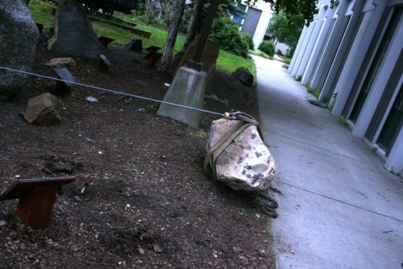 Rock with straps and cable attached, on dirt next to a concrete path and building