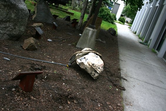Rock secured with a strap and cable on dirt ground next to a concrete path and building