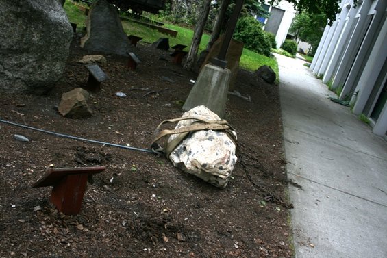 Rock with straps and chains on the ground near a concrete path and building