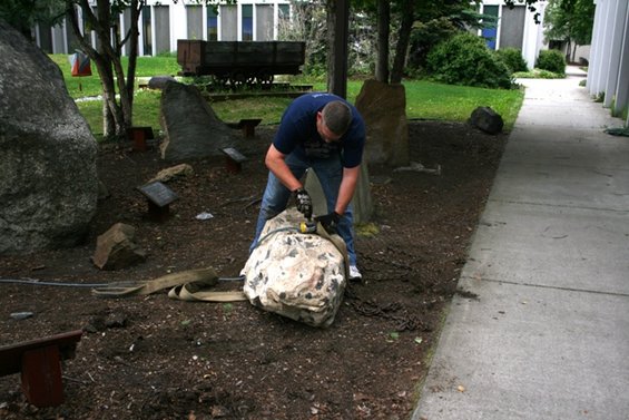 Man straps large rock for moving, with chains and a mining cart in background