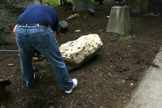 Man in blue shirt and jeans bends over a large rock in a park-like setting with chains on the ground