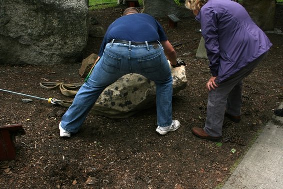 Two people are moving a large rock with straps and a metal bar in a rocky, wooded area