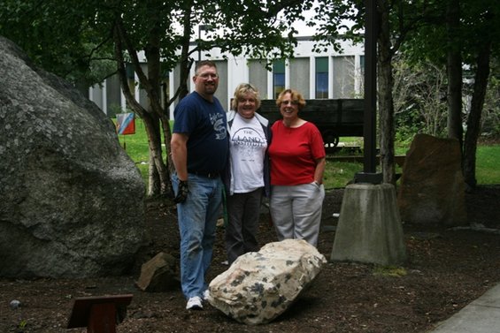 Three people stand in front of a building with large rocks and trees around them. One person wears a shirt that says 'The Land Institute'