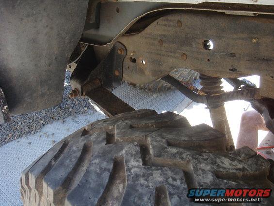Close-up of a large, knobby tire and rusty suspension components of a vehicle. A person's leg is visible in the background