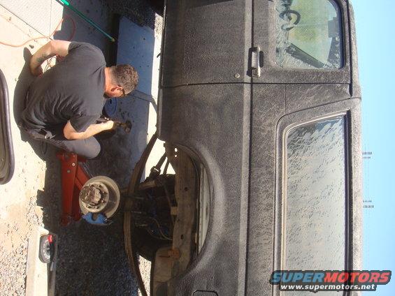 Man kneeling, working on the front axle of a dark-colored vehicle supported by a red jack