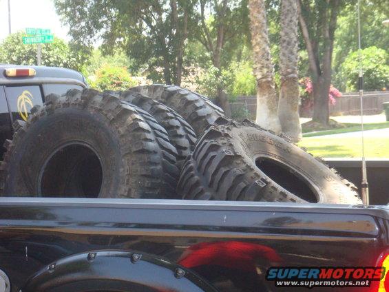 Truck bed loaded with four large, knobby off-road tires. A street sign is visible in the background