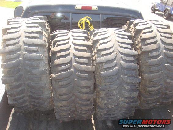 Four large off-road tires loaded in the bed of a pickup truck. A blue Jeep is visible in the background