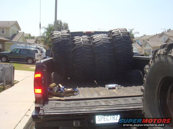 Pickup truck bed loaded with four large off-road tires. Another tire is visible on the right. A green SUV and a white van are parked in the background