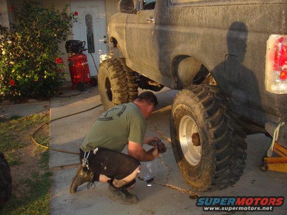 Man crouches to work on the rear axle of a lifted 1992 Ford Bronco. Large off-road tires are visible