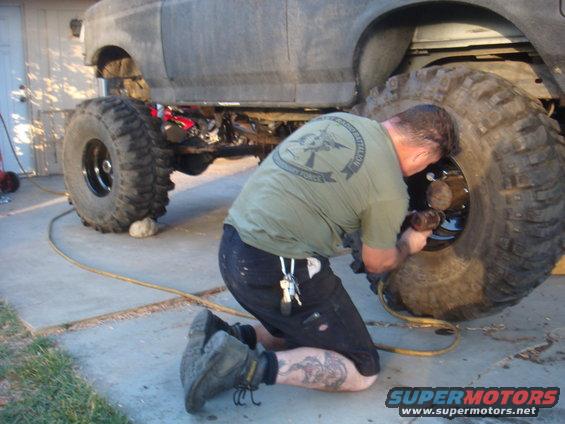 1992 Ford Bronco side view, man kneeling to work on a large tire, gray exterior, concrete driveway