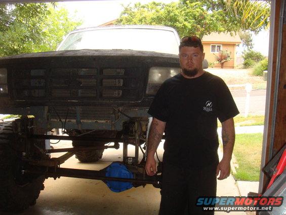 Man in black t-shirt stands before 1992 Ford Bronco front end and chassis. Truck is lifted with visible suspension and axle