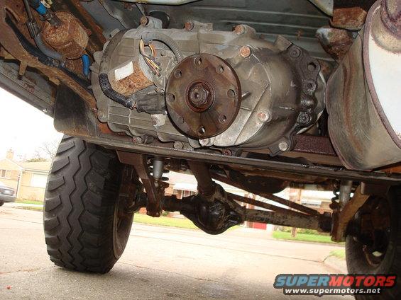 Transfer case and drivetrain of a vehicle, viewed from below. Rusty components and a large tire are visible