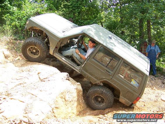 olive green SUV with large tires climbing over rocks, driver visible inside