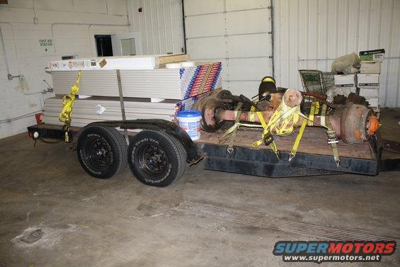 Trailer carrying two large axles and stacks of drywall sheets, secured with yellow straps