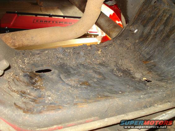 Undercarriage of a red vehicle with rust and dirt buildup, showing a pipe and part of a Craftsman jack