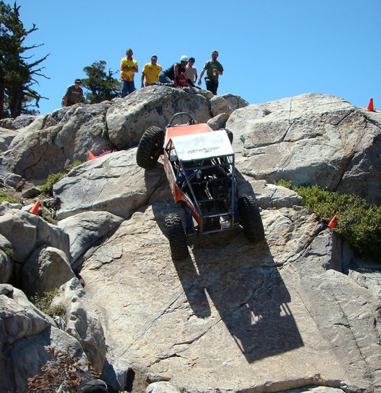 Orange off-road buggy with exposed engine and roll cage climbing a steep, rocky incline under a clear blue sky