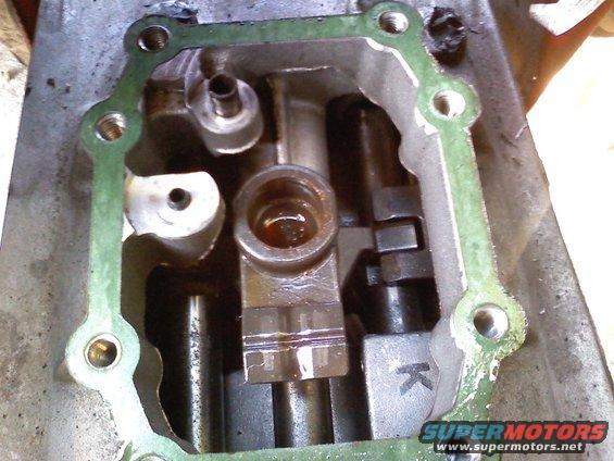Engine part with green gasket and visible internal components. A 'K' is stamped on one piece