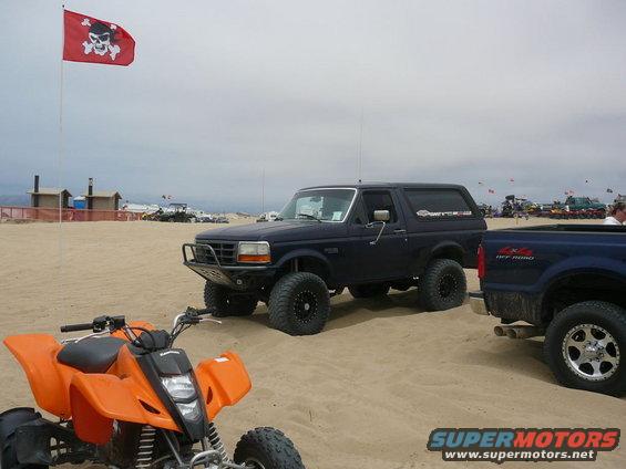 Dark blue Ford Bronco with aftermarket bumper and large tires parked on sand, orange ATV in foreground, blue pickup truck to the right