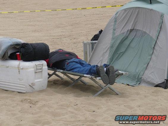 Person sleeping on a cot next to a cooler and a tent on a sandy surface