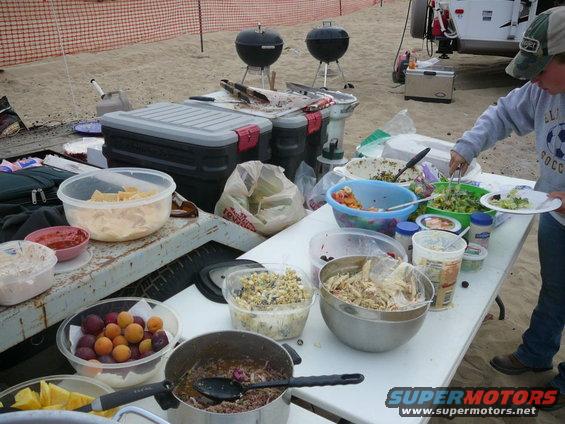 Outdoor picnic spread with food in bowls and containers, two grills, and a person serving salad